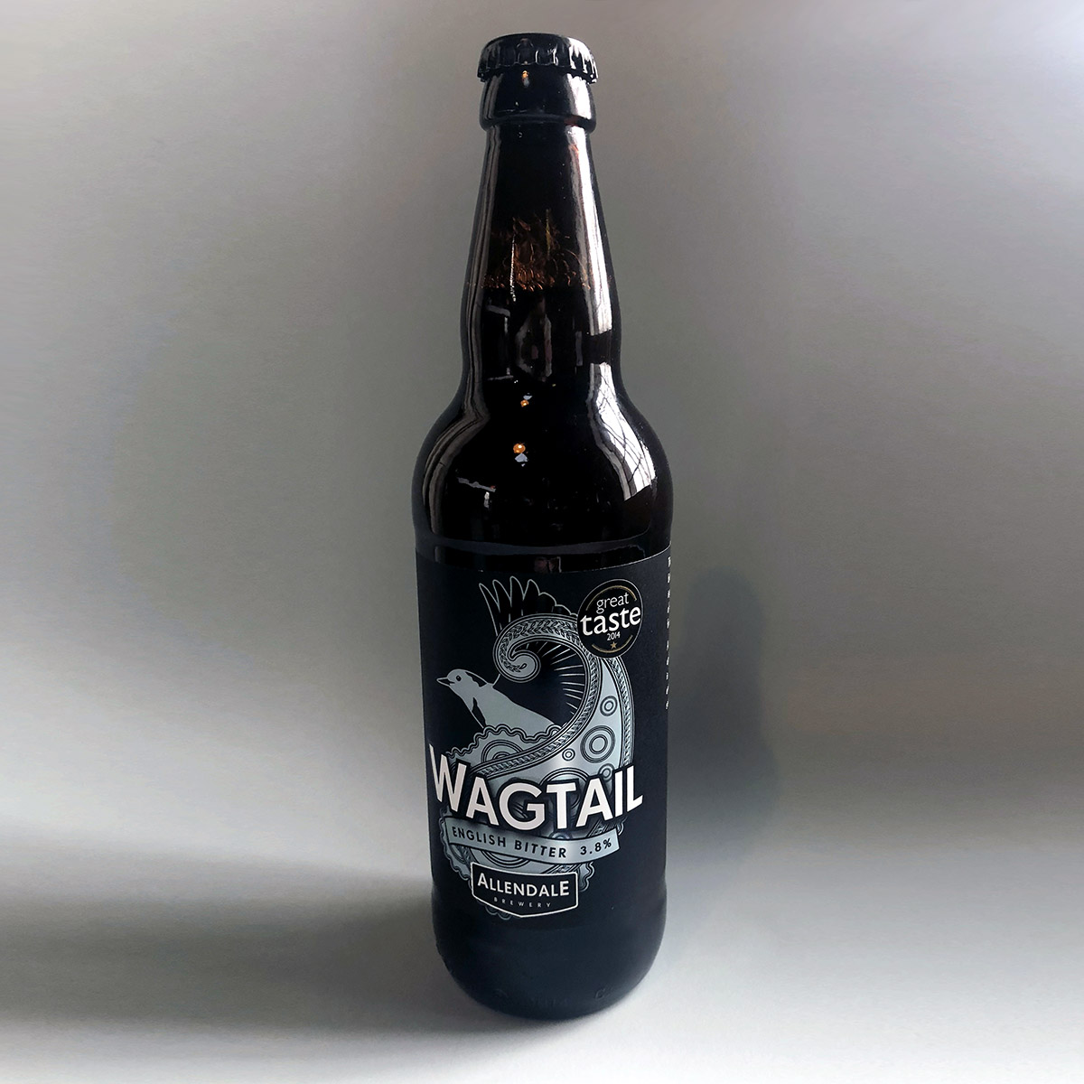 Image of a dark glass beer bottle on a grey background with the words 'WAGTAIL ENGLISH BITTER 3.8% ALLENDALE BREWERY' on it with an an image of a bird in a metallic blue and a 'great taste' award logo to the top right of the bottle label.