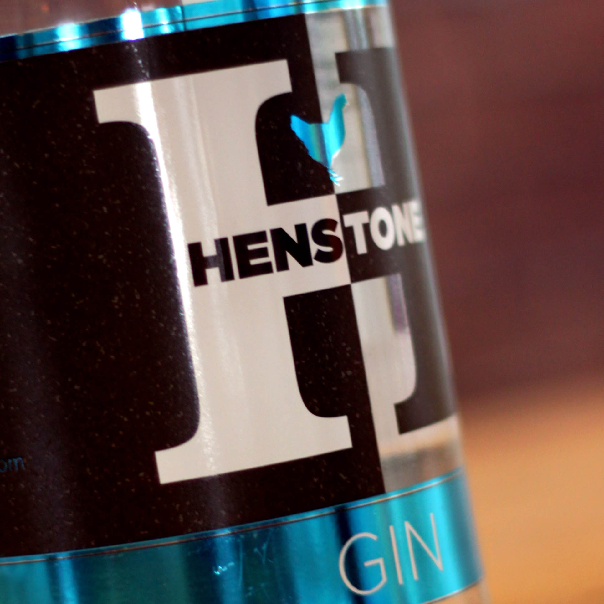 Image of a shiny metallic label fixed around a bottle with a large H as the central part of the image with the word 'HENSTONE' appearing across the centre of the H with a metallic 'electric' blue band underneath it with the word 'GIN' in white text