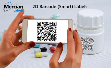 Two hands holding a smartphone with the image of a 2D (QR Code style) barcode on the phone. In the background is a pill bottle with the same 2D QR code style barcode on it and a few coloured pill capsules spread out on the table to the side of it.