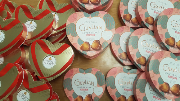 Multiple heart shaped boxes of two different brands of chocolates all piled up (offset - so creating an overlapping pattern) on top of each other on a desk.