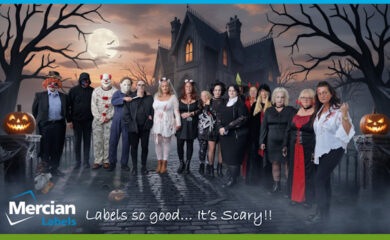 A group of Mercian Labels staff members dressed in Halloween costumes stand together in front of a spooky haunted house backdrop with a full moon, leafless trees, and glowing jack-o’-lanterns. The costumes include clowns, masked horror characters, witches, devils, gothic outfits, and zombie-like attire. The group is NOT smiling and posing for a fun company Halloween celebration. The Mercian Labels logo and the text “Labels so good… It’s Scary!!” appear at the bottom of the image.